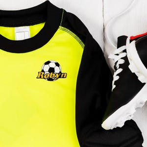 Puede incluir: Camiseta de f&uacute;tbol amarilla y negra con un bal&oacute;n de f&uacute;tbol negro y el nombre "Robyn" bordado en la parte delantera.