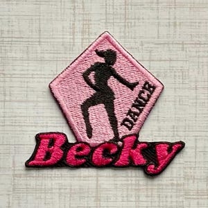 May include: A diamond-shaped embroidered patch featuring a light pink background with a black silhouette of a person dancing. The word DANCE is stitched in black on the right side, and the name Becky is embroidered in bright pink below the diamond.