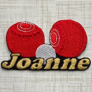 Könnte beinhalten: Ein gestickter Aufnäher mit zwei roten Rasen-Bocciakugeln und einer kleinen weißen Zielkugel über dem goldfarbenen Schriftzug Joanne. Das Design ist auf schwarzem Grund gehalten, wobei der Name in einer kursiven, fetten Gold-Schrift steht.