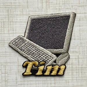 May include: An embroidered patch featuring a vintage computer monitor and keyboard in shades of silver and dark gray. Below the computer, the name Tim is stitched in bold, gold-colored thread with a black outline, creating a distinct graphic design.