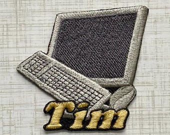 Könnte beinhalten: Ein gestickter Aufnäher mit einem Vintage-Computermonitor und einer Tastatur in Silber- und Dunkelgrautönen. Unter dem Computer ist der Name Tim in kräftigem, goldfarbenem Garn mit schwarzer Umrandung gestickt, was ein markantes Design ergibt.