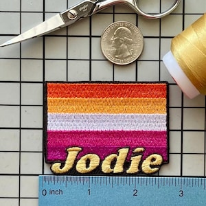 May include: A rectangular embroidered patch featuring horizontal stripes in shades of orange, yellow, white, and pink. The name Jodie is stitched in gold thread across the bottom. A ruler shows the patch is approximately 2.5 inches wide.