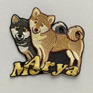 May include: An embroidered patch featuring two Shiba Inu dogs, one black and white and one tan and white, standing above the name Arya stitched in bold yellow letters with a dark outline against a solid black background.