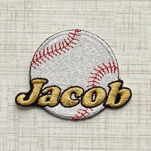 May include: A circular embroidered patch featuring a white baseball design with red stitching details. The name Jacob is embroidered across the center of the baseball in a bold, gold-colored font with a dark brown outline.