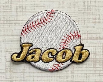 Könnte beinhalten: Ein runder, bestickter Aufnäher mit einem weißen Baseball-Design und roten Nahtdetails. Der Name Jacob ist in der Mitte des Baseballs in einer kräftigen, goldfarbenen Schrift mit dunkelbrauner Umrandung eingestickt.