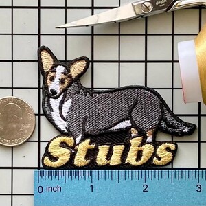May include: Embroidered patch featuring a gray and white corgi dog with the word "Stubs" in gold lettering. The patch is approximately 3 inches long and is placed on a grid background with a quarter coin for size comparison.