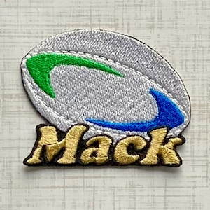 May include: An embroidered patch featuring a silver oval rugby ball shape with a green swoosh on the top left and a blue swoosh on the bottom right. Below the ball, the word Mack is stitched in gold thread with a dark brown outline.