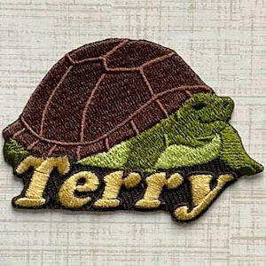May include: An embroidered patch featuring a green turtle with a dark brown shell. The turtle is depicted in profile, facing right. Below the turtle, the name Terry is stitched in large, light yellow, decorative, serif-style lettering.