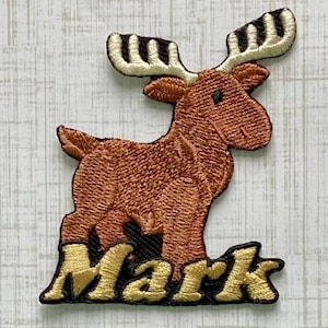 May include: An embroidered patch featuring a brown moose with cream-colored antlers. The moose stands above the name Mark, which is stitched in gold thread with a black outline. The entire design is bordered by a thin black embroidered edge.