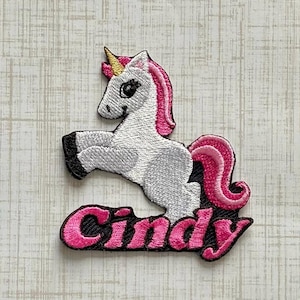 May include: An embroidered patch featuring a white unicorn with a pink mane and tail, a gold horn, and black hooves. The unicorn is positioned above the name Cindy, which is embroidered in bright pink script lettering with a black outline.