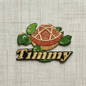 May include: An embroidered patch featuring a green turtle with a textured orange and cream shell. Below the turtle, the name Timmy is stitched in gold thread with a black outline, creating a decorative emblem with detailed thread work.