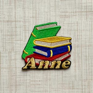 May include: An embroidered patch featuring a stack of four books in green, yellow, blue, and red with white pages. The name Anne is stitched in gold thread across the bottom of the stack in a bold, decorative font with a black outline.