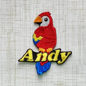 May include: An embroidered patch featuring a vibrant red parrot with a yellow and blue wing, a white face, and an orange beak. The parrot is perched on top of the name Andy, which is stitched in bright yellow letters with a black outline.