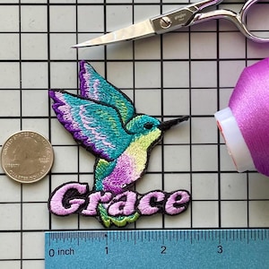May include: Embroidered hummingbird patch in shades of teal, purple, and yellow, with the name "Grace" in pink script. A quarter coin, scissors, and a spool of pink thread are also visible.