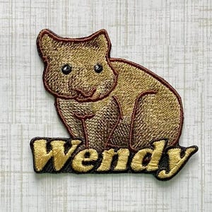 May include: An embroidered patch featuring a golden-brown quokka with dark brown outlines. Below the animal, the name Wendy is stitched in golden-yellow thread with a dark brown border, creating a cohesive design for this fabric applique.