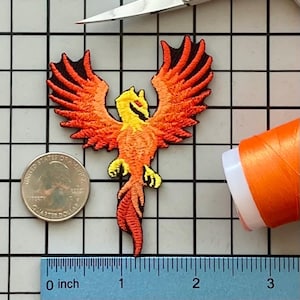 Può includere: Toppa ricamata raffigurante una fenice con le ali spiegate. L'uccello è raffigurato in tonalità di giallo, arancione e rosso, con dettagli neri. Una moneta da un quarto di dollaro e un righello sono accanto alla toppa. Una bobina di filo arancione e delle forbici sono anche nell'immagine.