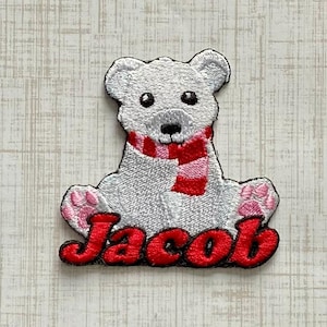 May include: An embroidered patch featuring a white polar bear cub with black eyes and nose, wearing a red and pink striped scarf. The bear has pink paw pads on its feet. The name Jacob is embroidered in bold red letters at the bottom.