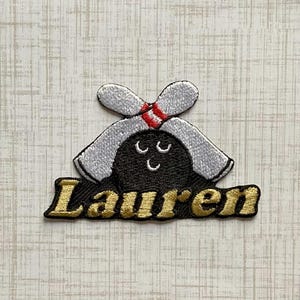 May include: An embroidered patch featuring a black bowling ball with a smiling face, topped with two crossed silver bowling pins with red stripes. The name Lauren is stitched in gold thread in a cursive font across the bottom of the patch.