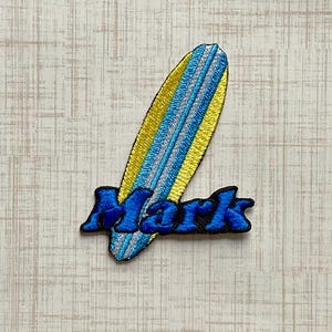 May include: An embroidered patch featuring a surfboard with yellow, light blue, and silver thread detailing. The word Mark is stitched across the lower portion of the surfboard in bold, dark blue thread with a thick black outline border.