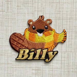 May include: An embroidered patch featuring a brown beaver wearing a yellow and orange striped scarf. The beaver has a black nose and white belly detail. Below the beaver, the name Billy is stitched in bold, gold-colored letters with a black outline.
