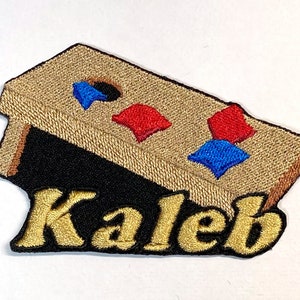 Puede incluir: Un parche bordado con un tablero de juego de madera marr&oacute;n con bolsas de frijoles rojas, azules y blancas. El nombre "Kaleb" est&aacute; bordado en oro debajo del tablero de juego.