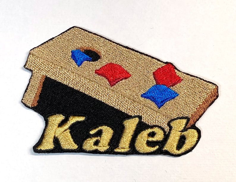 Puede incluir: Un parche bordado con un tablero de juego de madera marr&oacute;n con bolsas de frijoles rojas, azules y blancas. El nombre "Kaleb" est&aacute; bordado en oro debajo del tablero de juego.