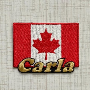 May include: A rectangular embroidered patch featuring the Canadian flag design with a red maple leaf on a white center panel flanked by red side panels. The name Carla is embroidered in gold thread with a black outline across the bottom.