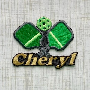 May include: An embroidered patch featuring two green pickleball paddles crossed behind a lime green pickleball. The name Cheryl is stitched in gold thread with a black outline across the bottom of the design, creating a cohesive sports-themed emblem.