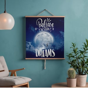 Puede incluir: Un tapiz rectangular que muestra un cielo nocturno azul oscuro con una gran luna llena y nubes. El texto dice Believe IN YOUR DREAMS con una pequeña ilustración de una figura de palo. La impresión se sujeta con dos barras de madera horizontales.