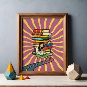 May include: A framed wall art print featuring a person sitting cross-legged and reading a red book in front of a large stack of colorful books. The background displays a yellow and magenta sunburst pattern, with a blue road leading toward the person.
