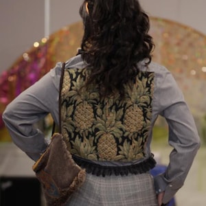 May include: A light blue long-sleeve button-down shirt with a black and gold pineapple-patterned back panel, paired with grey plaid trousers featuring black lace trim at the hem and a small brown shoulder bag with a fur-like texture.