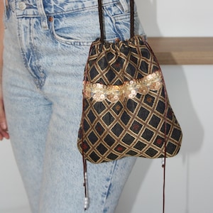 May include: A black drawstring bag with a diamond pattern in gold and red. The bag features a band of gold sequins and brown drawstrings with pearl accents. The bag is held by a person wearing blue jeans.