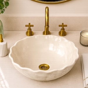 May include: A white ceramic vessel sink with a scalloped, wavy rim and a smooth, glossy finish. The sink is paired with a brushed gold faucet featuring two cross-handle controls and a matching gold drain assembly centered in the basin.