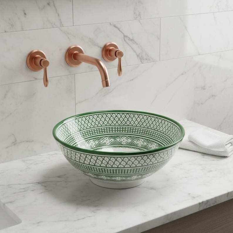 May include: A decorative ceramic sink with a green and white geometric pattern sits on a marble countertop. Above, copper-colored faucet fixtures are mounted on a white tiled wall. A white hand towel is placed to the right of the sink.