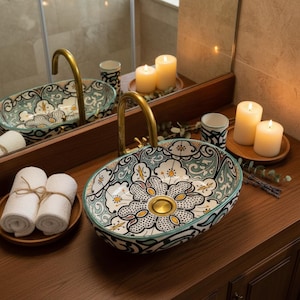 May include: An oval ceramic bathroom sink with a floral pattern in white, teal, and yellow. The sink has a gold tap and drain. Rolled white towels and candles are on the wooden counter. A mirror reflects the sink and candles.