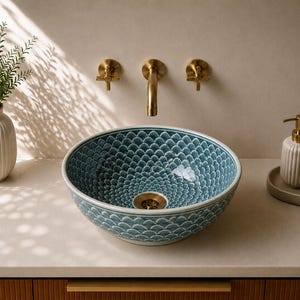 Puede incluir: Un lavabo de baño moderno con una cuenca estampada en azul y blanco. Se combina con grifos dorados y un dispensador de jabón y una taza a juego. Un jarrón blanco con vegetación está a la izquierda.