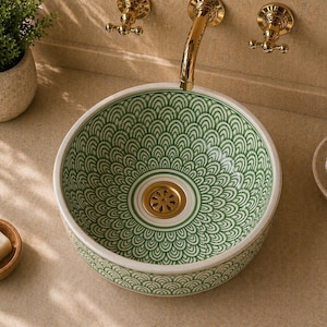 May include: A decorative, round ceramic sink with a green and white patterned design. The sink has a gold-colored drain and faucet. A small wooden bowl with soap and a small potted plant are nearby.