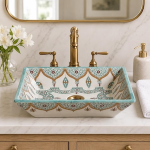 May include: A rectangular ceramic vessel sink featuring a white base with intricate teal, gold, and maroon floral patterns along the interior and exterior rims. The sink is paired with a brushed brass faucet and two matching lever handles.