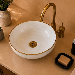 May include: A round white ceramic vessel sink with a gold-toned rim and a matching gold-toned drain. The sink is paired with a tall, curved gold-toned tap. The basin features a smooth, glossy finish and sits on a flat, light-coloured countertop.