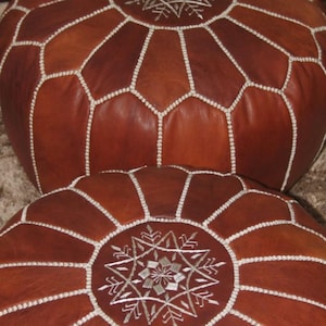 Op de afbeelding: Twee bruine Marokkaanse poefs van leer. De poefs zijn rond met een geometrisch patroon van gestikte witte lijnen. De bovenkant van elke poef heeft een decoratief zilverkleurig geborduurd ontwerp. Het leer heeft een rijke, warme tint.