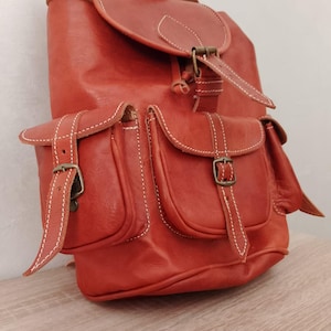 Może przedstawiać: Ciemnopomarańczowy skórzany plecak z dwiema przednimi kieszeniami i klapą. Torba posiada białe przeszycia i mosiężne klamry. Posiada górny uchwyt i regulowane paski.