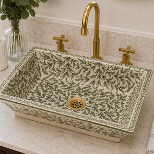 Puede incluir: Un lavabo rectangular de cerámica con un patrón de hojas verdes pintado a mano sobre un fondo crema. El cuenco tiene un borde ancho y plano con una cenefa decorativa y un desagüe central en tono latón, acompañado de un grifo de latón curvo.