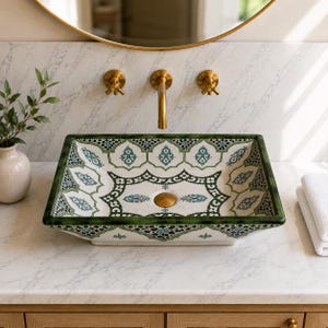 May include: A rectangular ceramic vessel sink featuring a white base with intricate green and teal geometric patterns. The sink has a dark green rim and a central brass drain cover, positioned on a white marble countertop with a brass faucet.