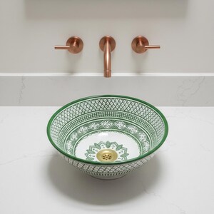 May include: A decorative ceramic sink with a green and white geometric pattern. The sink has a gold drain and is set on a white countertop. Copper-colored faucet fixtures are mounted on the wall above the sink.