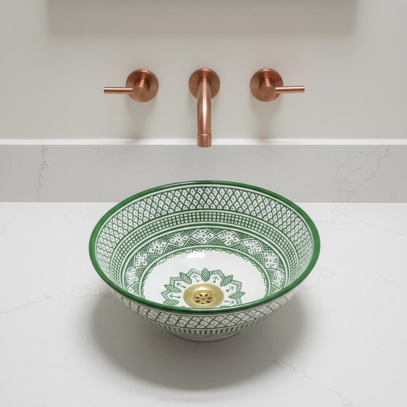 May include: A decorative ceramic sink with a green and white geometric pattern. The sink has a gold drain and is set on a white countertop. Copper-colored faucet fixtures are mounted on the wall above the sink.
