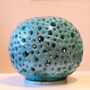 Può includere: Una sfera di ceramica turchese con numerosi fori di dimensioni irregolari. L'oggetto decorativo ha una finitura lucida e poggia su una piccola base circolare. Il colore è una miscela di blu e verde, creando un aspetto unico e strutturato.