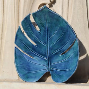 Puede incluir: Una pieza decorativa de cerámica con forma de hoja en tonos azules. La hoja tiene un acabado brillante y vetas detalladas, con secciones recortadas en los bordes. El fondo es una superficie texturizada de color blanquecino.