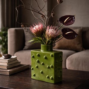 Può includere: Un vaso quadrato in ceramica verde con superficie strutturata, pieno di fiori rosa e bianchi, anthurium viola scuro e rami secchi. Il vaso è su un tavolo di legno scuro, con un divano sullo sfondo.