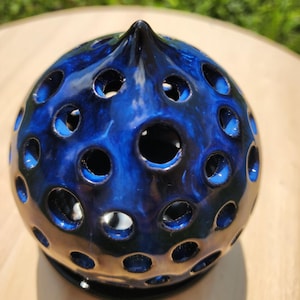 Op de afbeelding: Een keramische, bolvormige kandelaar met een glanzend, donkerblauw glazuur. Het oppervlak is geperforeerd met talrijke cirkelvormige openingen. De bovenkant heeft een puntige punt. De kandelaar staat op een kleine, zwarte basis.