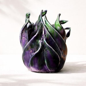 May include: A ceramic vase with a unique, organic design. The vase features a blend of deep purple and green hues, with a glossy finish. The form is reminiscent of stylized petals or flames, creating a sculptural effect. The vase is a decorative item.